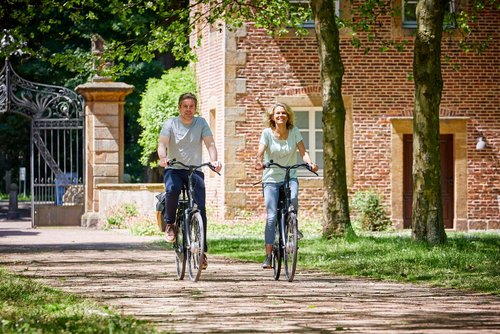 Zwei Personen fahren Fahrrad auf einem Weg, der von einem prunkvollen Tor und einem Backsteingebäude wegführt.