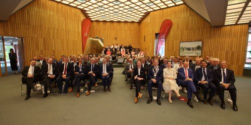 In einem Sitzungssaal sitzen zahlreiche Personen in mehreren Stuhlreihen und schauen in die Kamera. Zwei rote Banner tragen die Aufschrift "Kreis Coesfeld".
