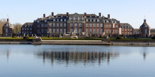 Ein barockes Schloss mit symmetrischem Aufbau ist hinter einem Gewässer zu sehen.
