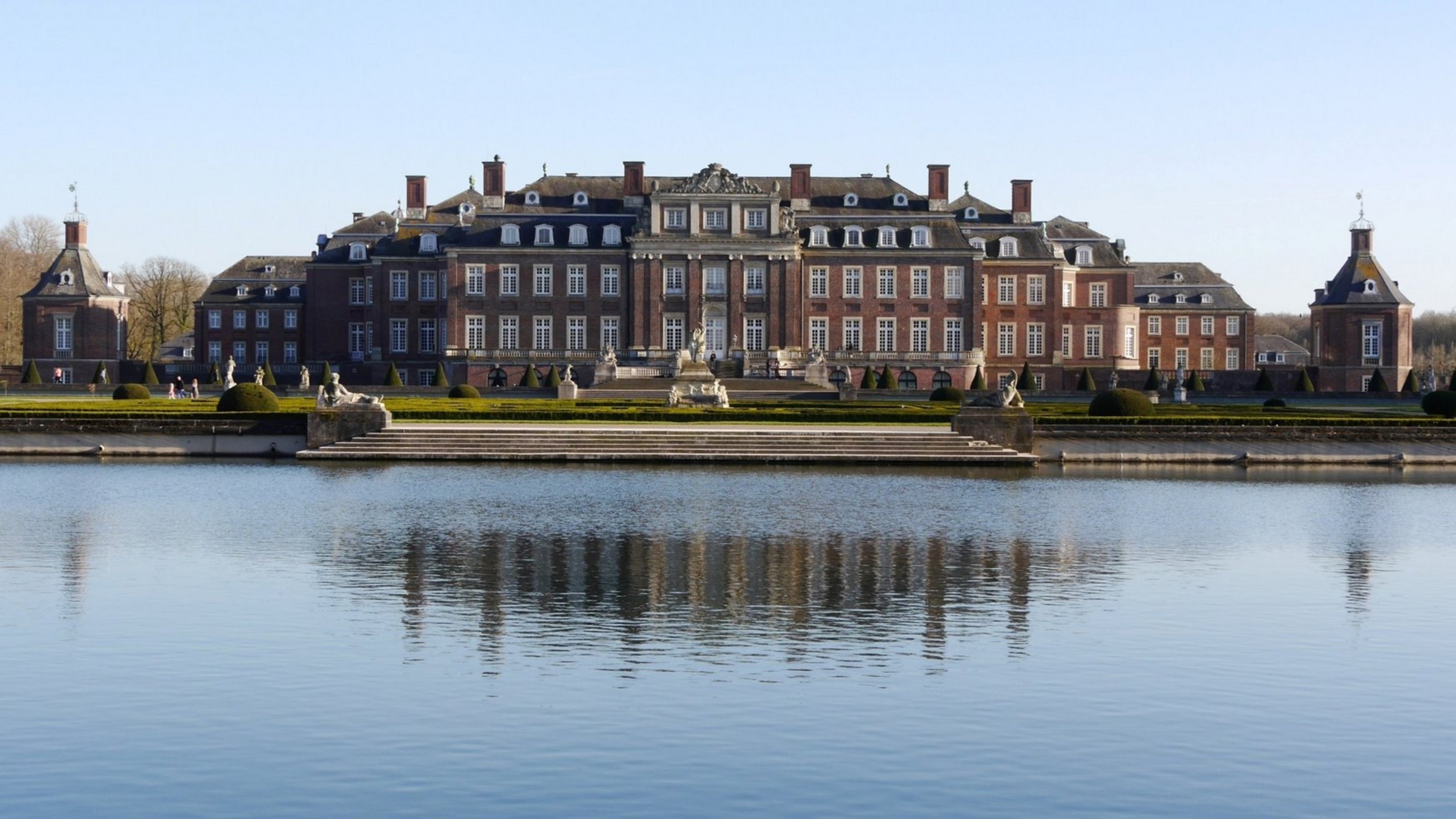 Ein barockes Schloss mit symmetrischem Aufbau ist hinter einem Gewässer zu sehen.