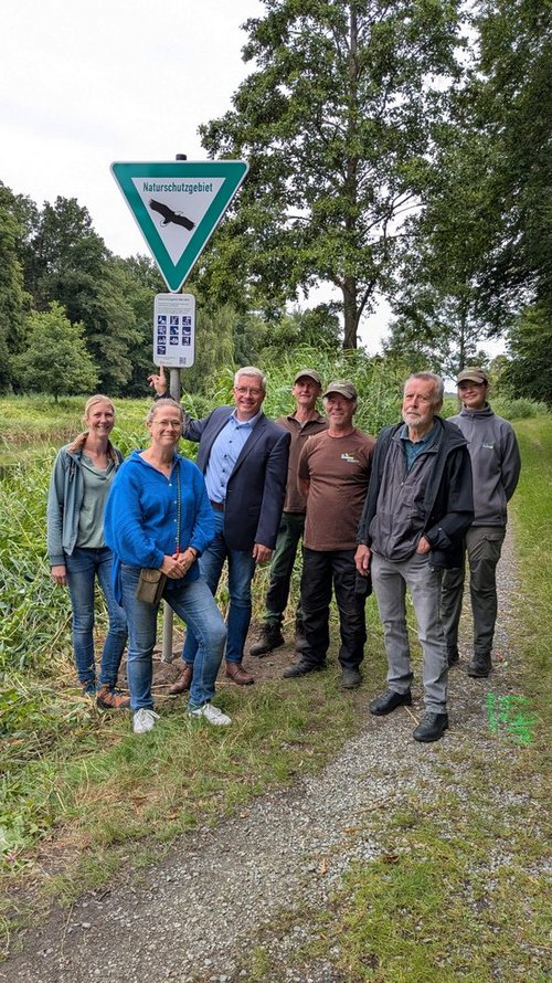 Eine Gruppe von sieben Personen steht auf einem Weg neben einem Naturschutzgebietsschild mit Piktogrammen und QR-Code. Im Hintergrund sind Bäume, Sträucher und ein Gewässer zu sehen. Die Gruppe lächelt in die Kamera; eine Person zeigt auf das Schild.