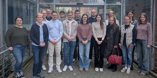 Eine Gruppe steht in zwei Reihen vor dem Eingangsbereich eines Gebäudes mit der Aufschrift "Gesundheitsamt". Die Gruppe besteht aus 16 Personen.