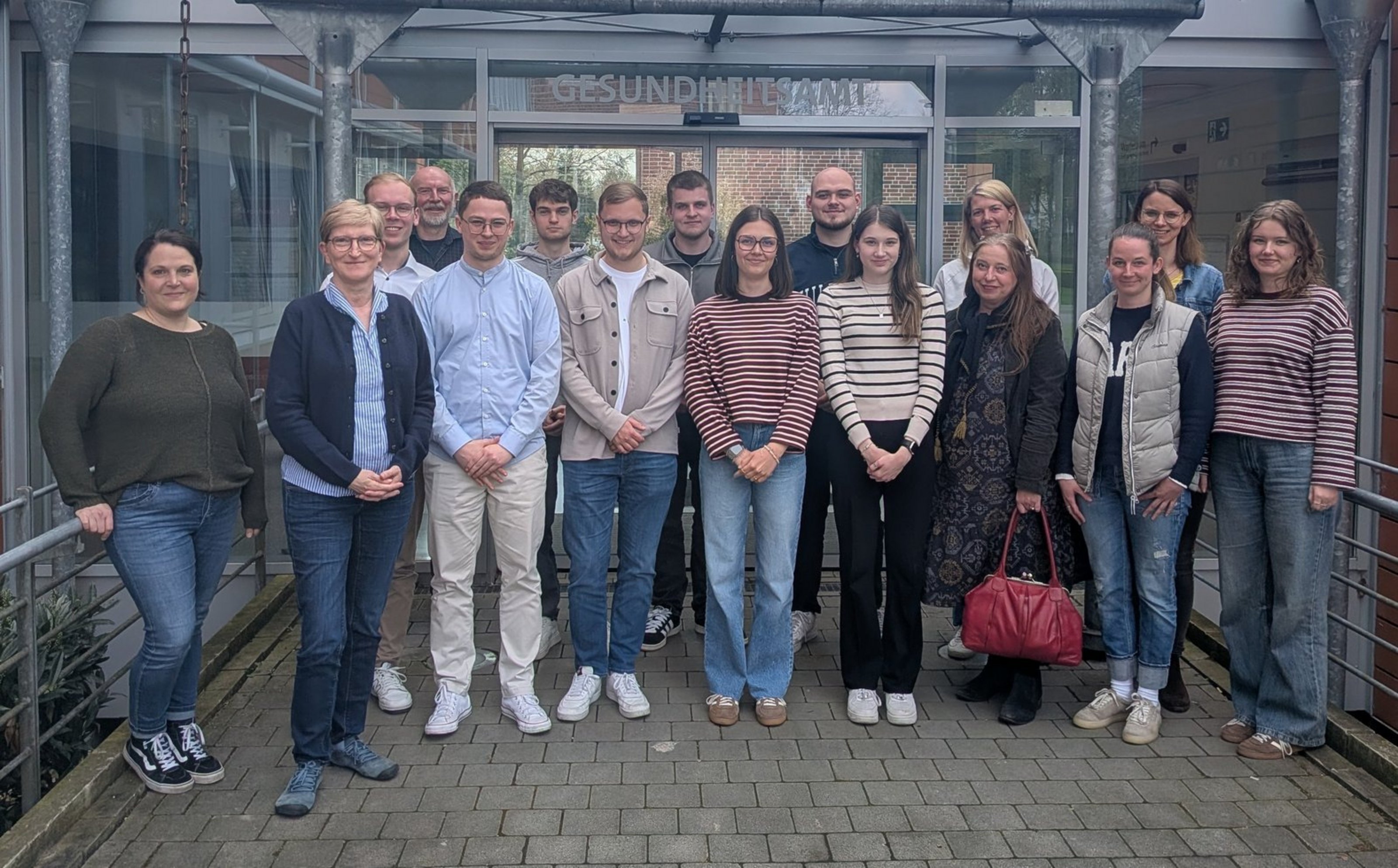 Eine Gruppe steht in zwei Reihen vor dem Eingangsbereich eines Gebäudes mit der Aufschrift "Gesundheitsamt". Die Gruppe besteht aus 16 Personen.