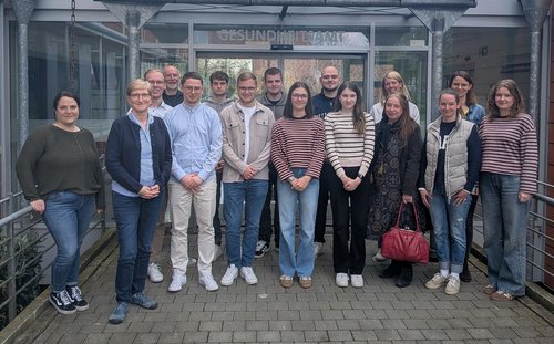 Eine Gruppe steht in zwei Reihen vor dem Eingangsbereich eines Gebäudes mit der Aufschrift "Gesundheitsamt". Die Gruppe besteht aus 16 Personen.