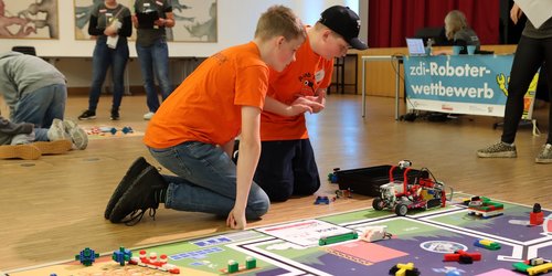 Zwei Jungen hocken vor einem Spielfeld. Auf dem Spielfeld befinden sich Klemmsteine und ein Roboter. Im Hintergrund ist ein Plakat mit der Aufschrift "zdi-Roboter-Wettbewerb" und zwei Frauen stehen weiter hinten. Die Aufnahme wurde in einem großen Raum mit Parkettboden aufgenommen..