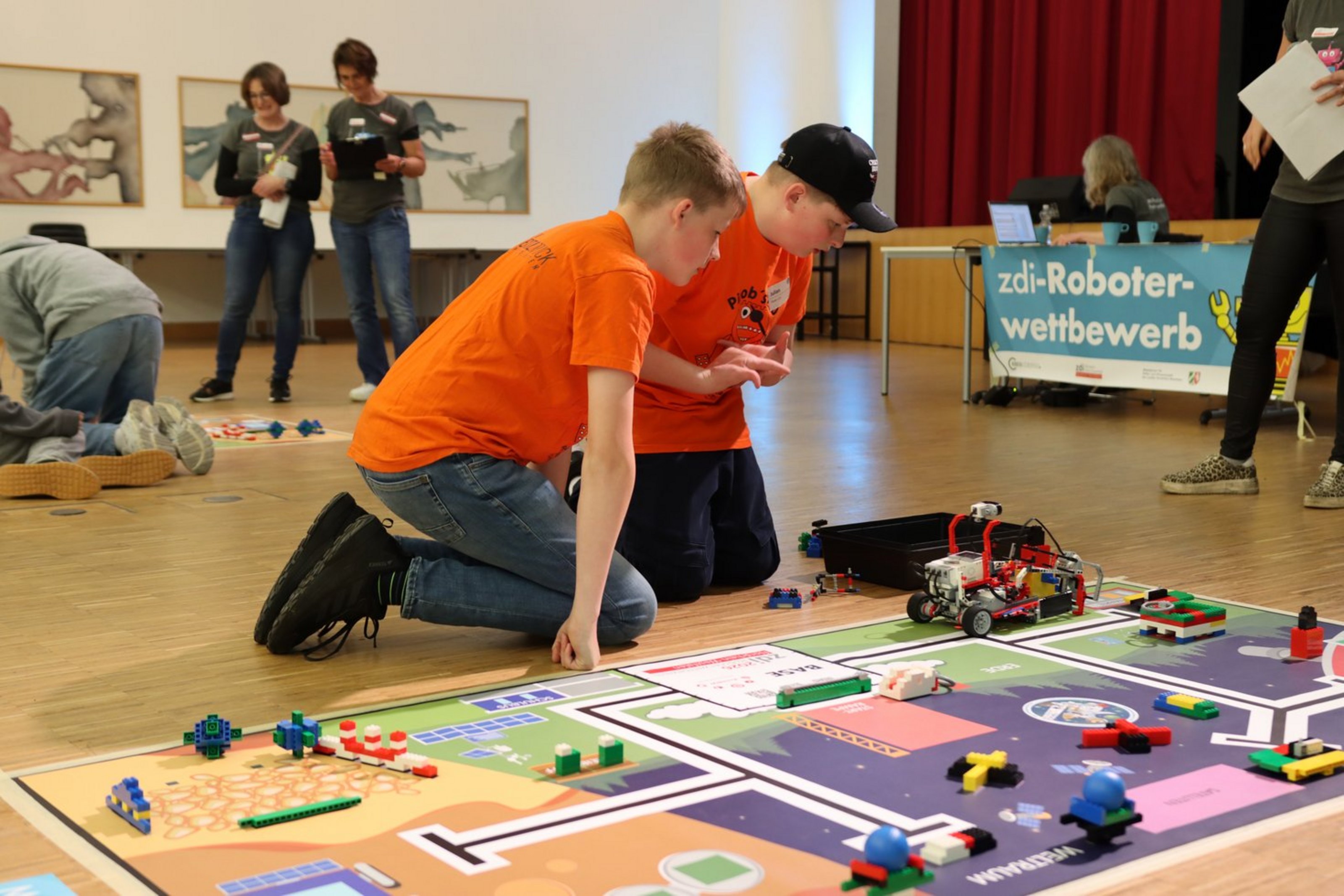 Zwei Jungen hocken vor einem Spielfeld. Auf dem Spielfeld befinden sich Klemmsteine und ein Roboter. Im Hintergrund ist ein Plakat mit der Aufschrift "zdi-Roboter-Wettbewerb" und zwei Frauen stehen weiter hinten. Die Aufnahme wurde in einem großen Raum mit Parkettboden aufgenommen..