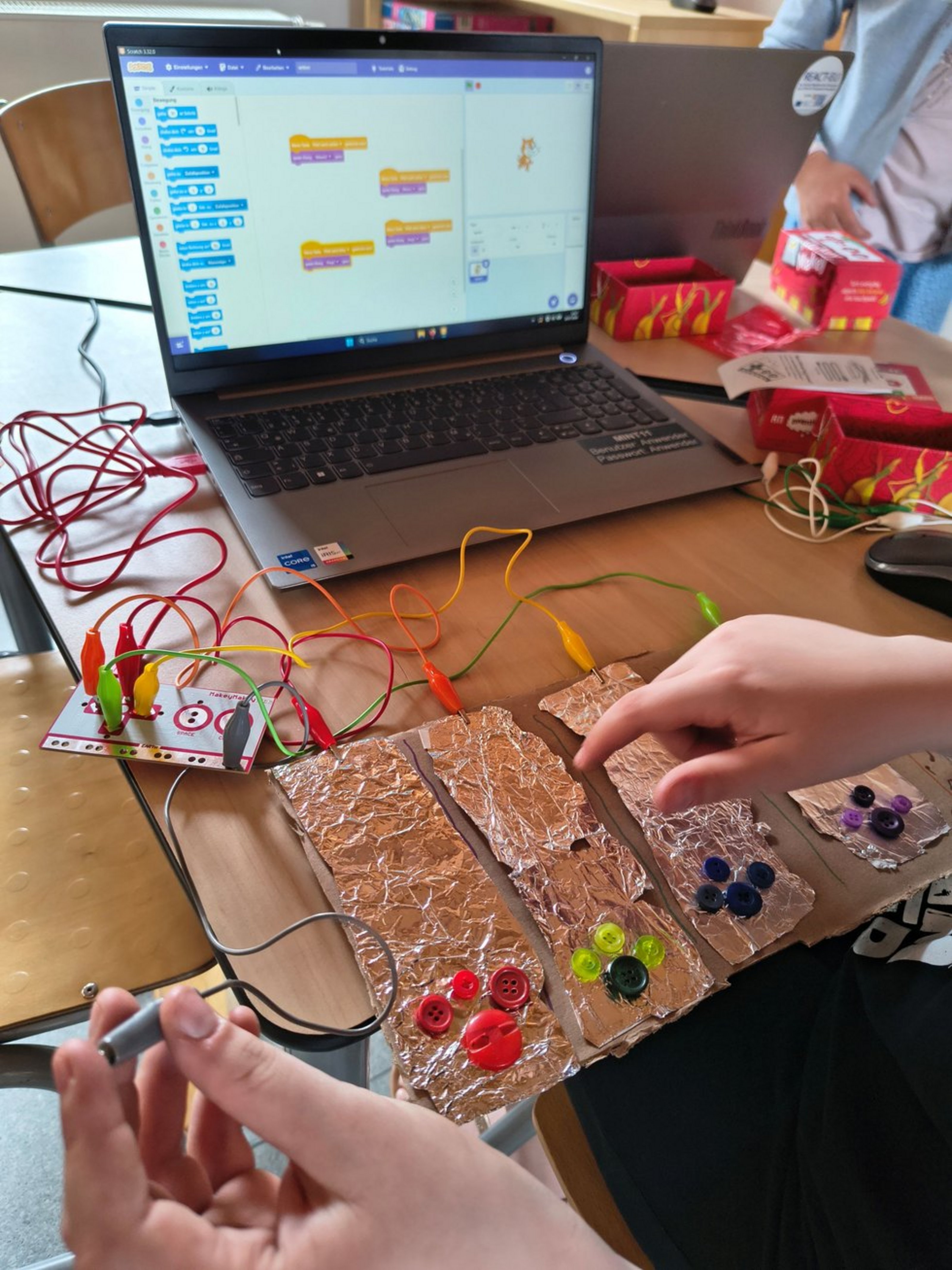 Auf einem Tisch steht ein aufgeklappter Laptop, vor dem ein Stück bemalte Pappe liegt. An der Pappe sind Stromkabel befestigt, die zum Laptop führen.
