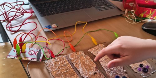 Auf einem Tisch steht ein aufgeklappter Laptop, vor dem ein Stück bemalte Pappe liegt. An der Pappe sind Stromkabel befestigt, die zum Laptop führen.