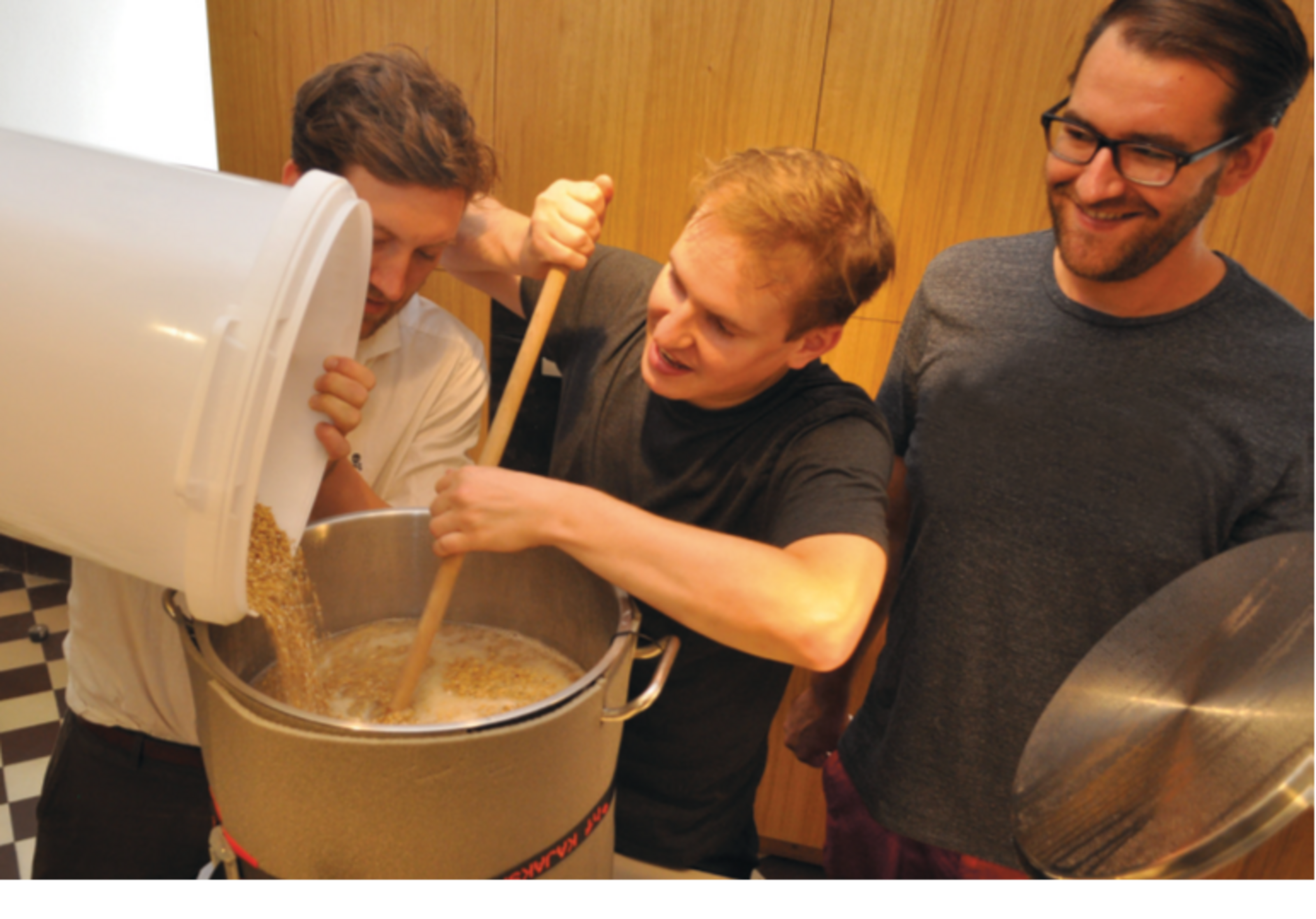 Bierbrauen in der Burgküche (Foto: Kreis Coesfeld, Markus Kleymann). Drei Männer stehen um einen Topf, in dem sich eine milchige Flüssigkeit befindet. Einer rührt mit einem Holzstab darin. Ein zweiter schüttet Körner in den Topf.