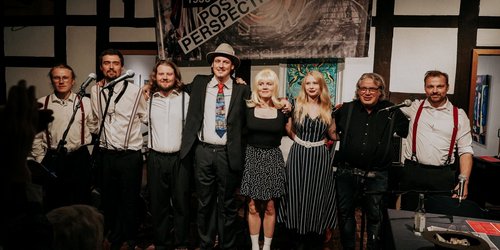 Acht Personen stehen untergeharkt vor einer Wand aus Fachwerk. Sie tragen Hemden und Hosenträger bzw. Kleider.