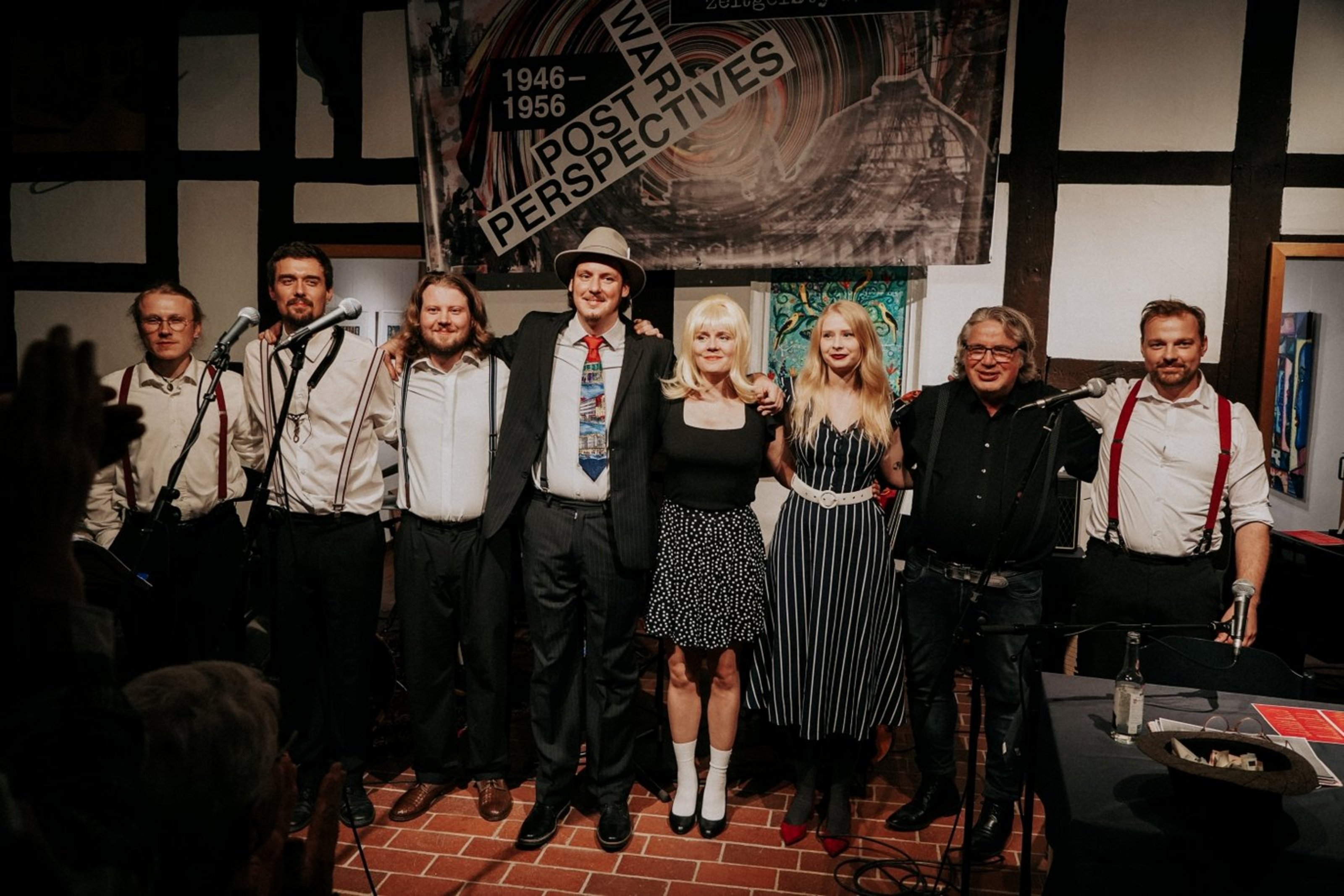 Acht Personen stehen untergeharkt vor einer Wand aus Fachwerk. Sie tragen Hemden und Hosenträger bzw. Kleider.