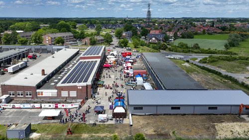 Luftaufnahme von einem betrieblichen Gelände mit zwei Hallen. Auf dem Zwischenhof sind Pavillions und Fahrzeuge aufgestellt. Viele Menschen sind dort zu sehen.