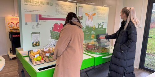 Zwei Personen stehen vor einer Schautafel mit der Aufschrift "Immer  lecker, immer frisch".