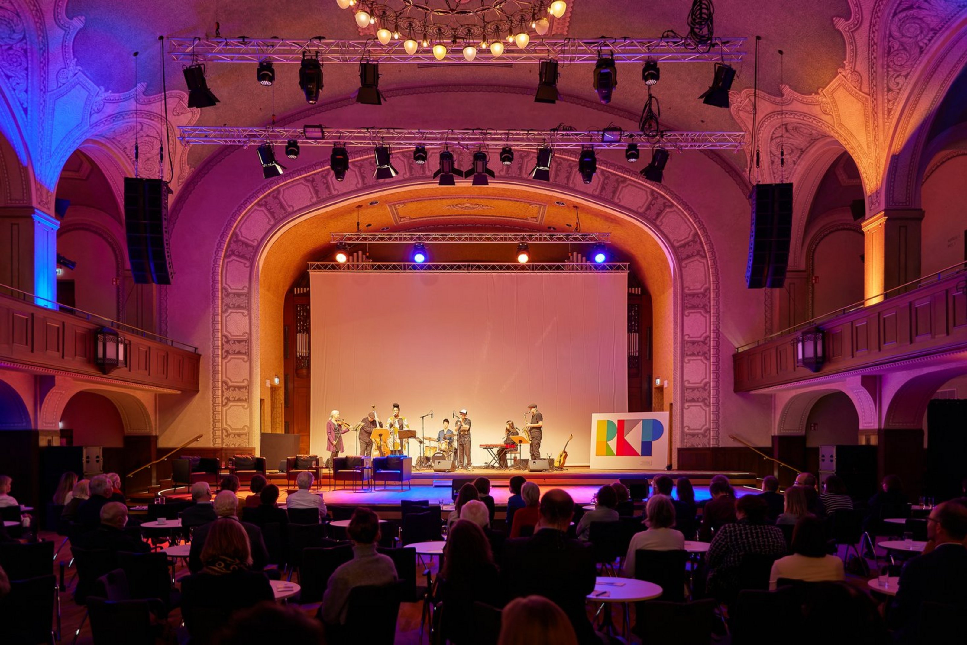 Ein Konzertsaal mit Publikum in rotem Licht ist zu sehen. Auf der Bühne steht eine Band bestehend aus sieben Personen.