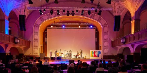 Ein Konzertsaal mit Publikum in rotem Licht ist zu sehen. Auf der Bühne steht eine Band bestehend aus sieben Personen.
