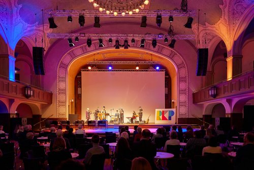 Ein Konzertsaal mit Publikum in rotem Licht ist zu sehen. Auf der Bühne steht eine Band bestehend aus sieben Personen.