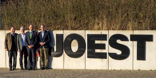 Vier Männer in Anzügen stehen auf der linken Bildseite vor einer Mauer. Neben Ihn der Schriftzug "JOEST".