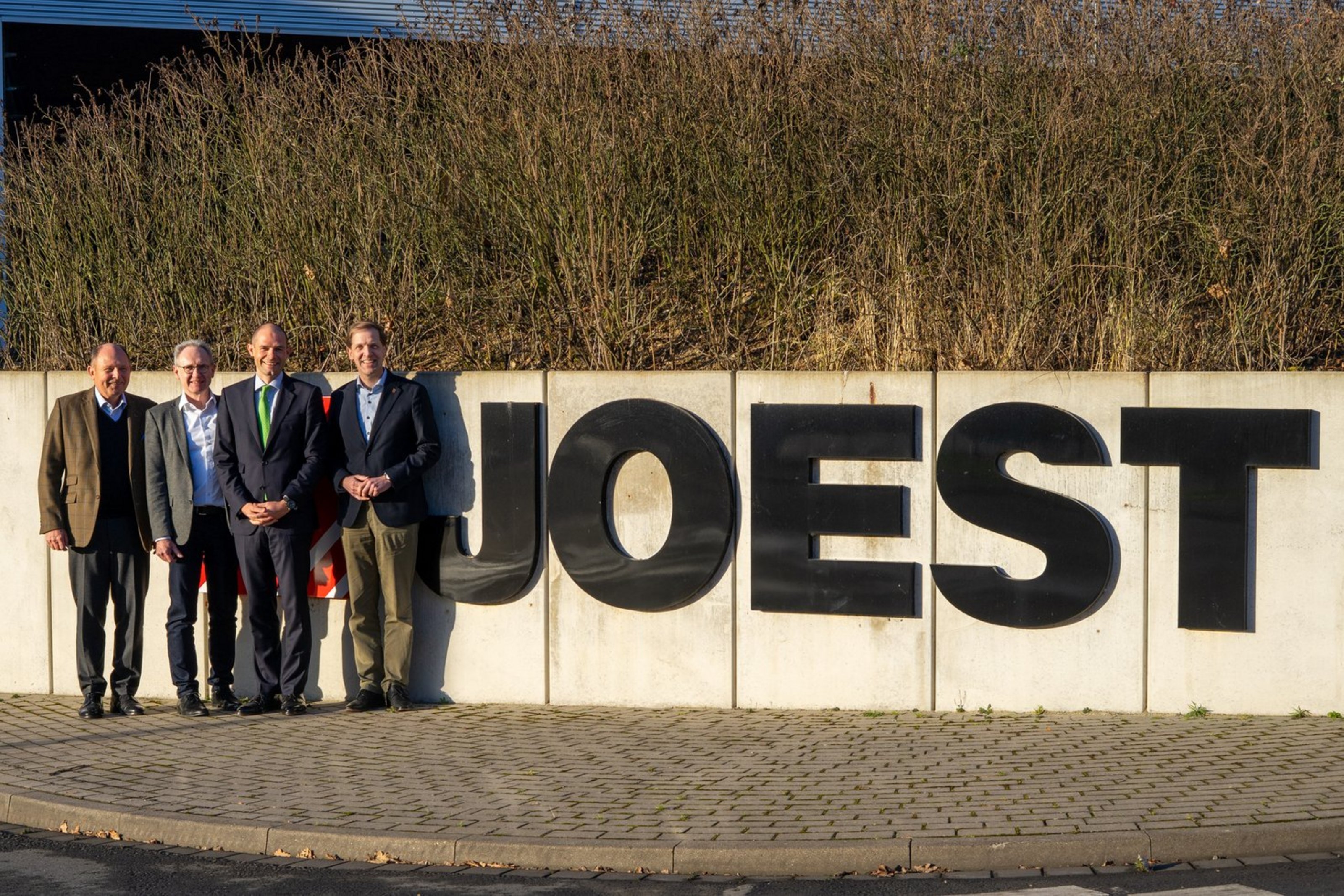 Vier Männer in Anzügen stehen auf der linken Bildseite vor einer Mauer. Neben Ihn der Schriftzug "JOEST".