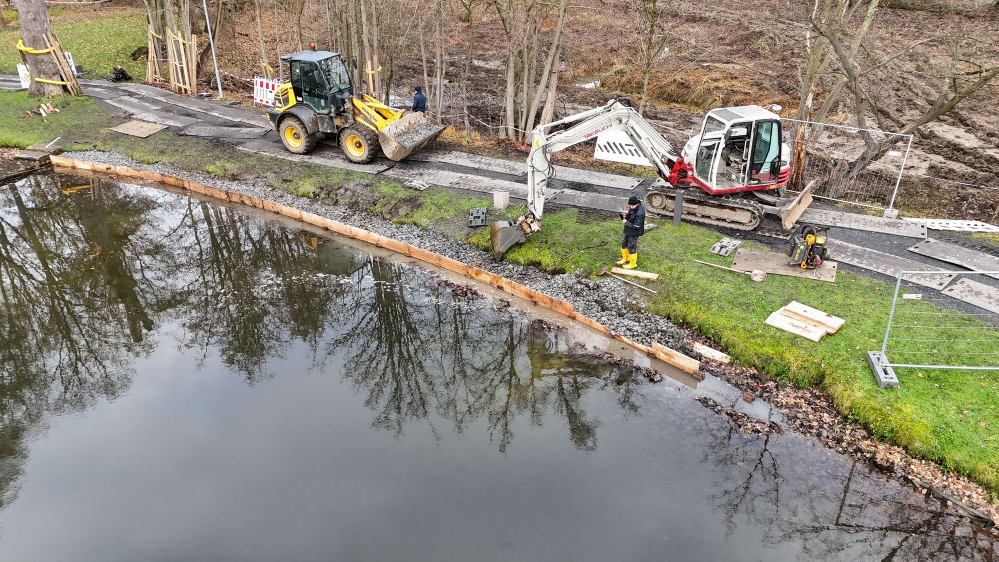 Luftaufnahme von einer Gewässerkante, an der Baumaterial liegt sowie zwei Bagger  zu sehen sind.