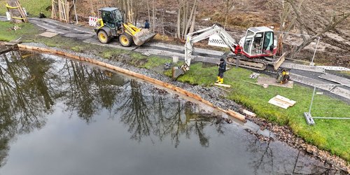 Luftaufnahme von einer Gewässerkante, an der Baumaterial liegt sowie zwei Bagger  zu sehen sind.