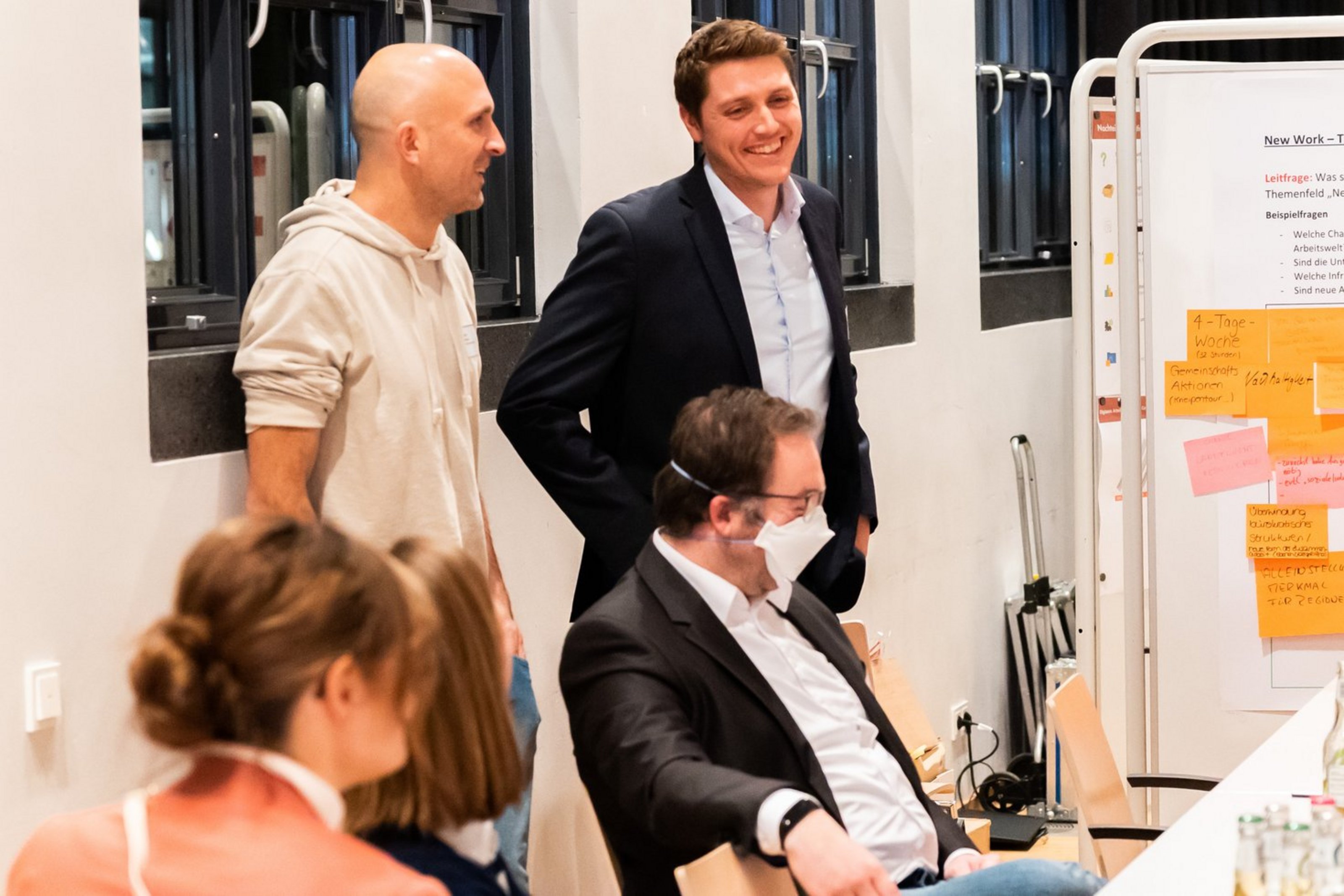 Vier Personen in einem Besprechungsraum, zwei sitzen, zwei stehen, einer trägt einen Mund-Nasen-Schutz