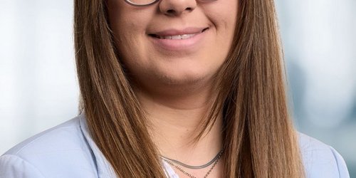 Eine Porträt zeigt eine Frau mit langen Haaren und Brille. Sie trägt einen Blazer.