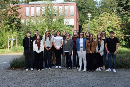 16 Personen stehen in zwei Reihen. Im Hintergrund sind ein Backsteingebäude und Büsche zu sehen.
