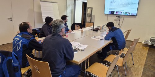 Fünf Männer in blauer Arbeitskleidung sitzen an einem Gruppentisch und schauen zur Wand auf eine digitale Tafel. Auf dem Tisch liegen Schreibutensilien und Bücher.
