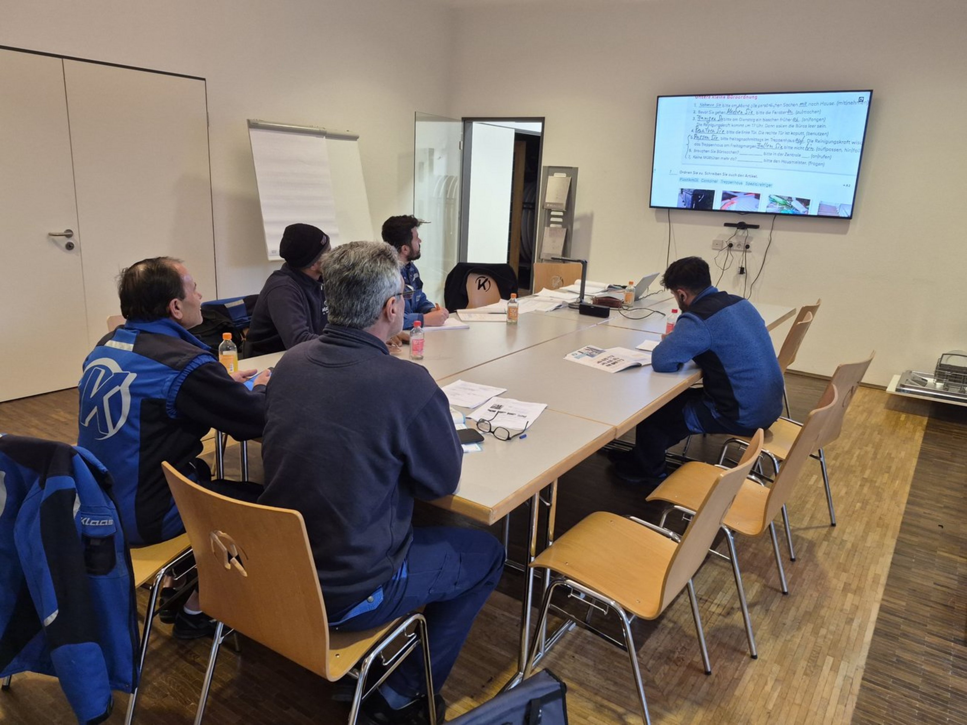 Fünf Männer in blauer Arbeitskleidung sitzen an einem Gruppentisch und schauen zur Wand auf eine digitale Tafel. Auf dem Tisch liegen Schreibutensilien und Bücher.