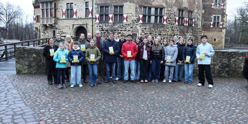 Eine Gruppe aus Jugendlichen und Erwachsenen steht vor einer Mauer und halten jeweils ein kleines grünes Büchlein in der Hand. Dahinter ist eine Burg.
