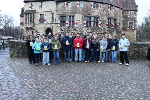 Eine Gruppe aus Jugendlichen und Erwachsenen steht vor einer Mauer und halten jeweils ein kleines grünes Büchlein in der Hand. Dahinter ist eine Burg.