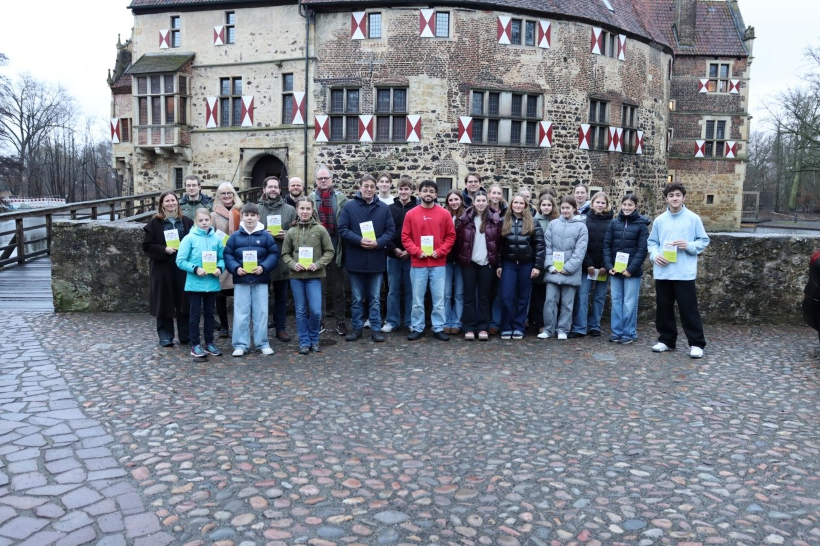 Eine Gruppe aus Jugendlichen und Erwachsenen steht vor einer Mauer und halten jeweils ein kleines grünes Büchlein in der Hand. Dahinter ist eine Burg.
