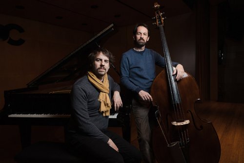 Zwei Musiker posieren in einem Konzertsaal – einer sitzt am Klavier, der andere steht mit einem Kontrabass.