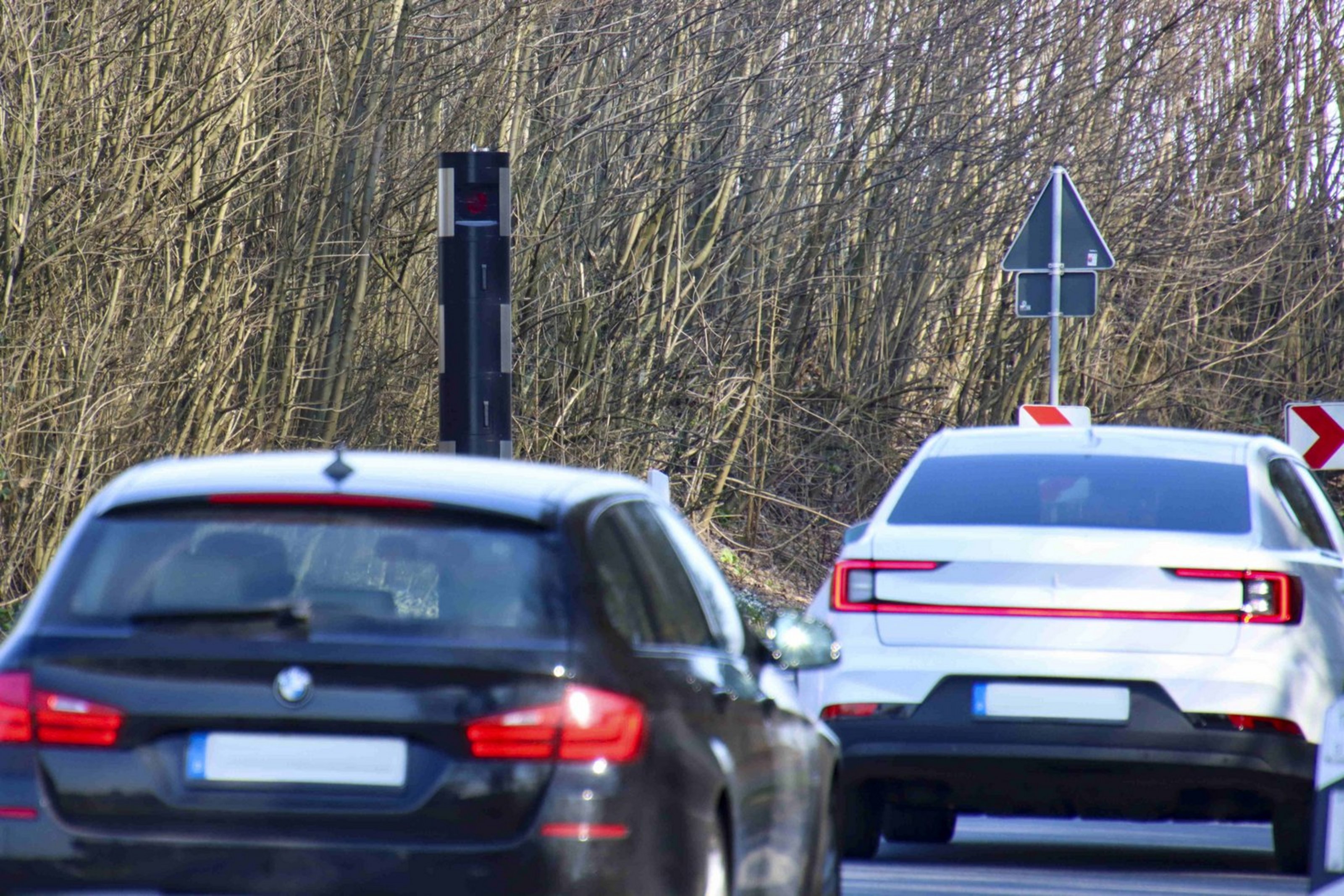 Auch in der kommenden Woche wird der Kreis an bestimmten Stellen Geschwindigkeitskontrollen durchführen (Bildquelle: Kreis Coesfeld). Zwei Autos fahren auf einer Landstraße an einem stationären Blitzer vorbei. Der schwarze Blitzer steht am Fahrbahnrand vor einem Waldstück und zeigt ein rotes Lichtsignal an. Im Hintergrund sind Verkehrszeichen zu sehen, darunter ein Vorfahrt-achten-Schild und rot-weiße Richtungspfeile. Die Szene spielt sich an einer leichten Kurve bei Tageslicht ab.