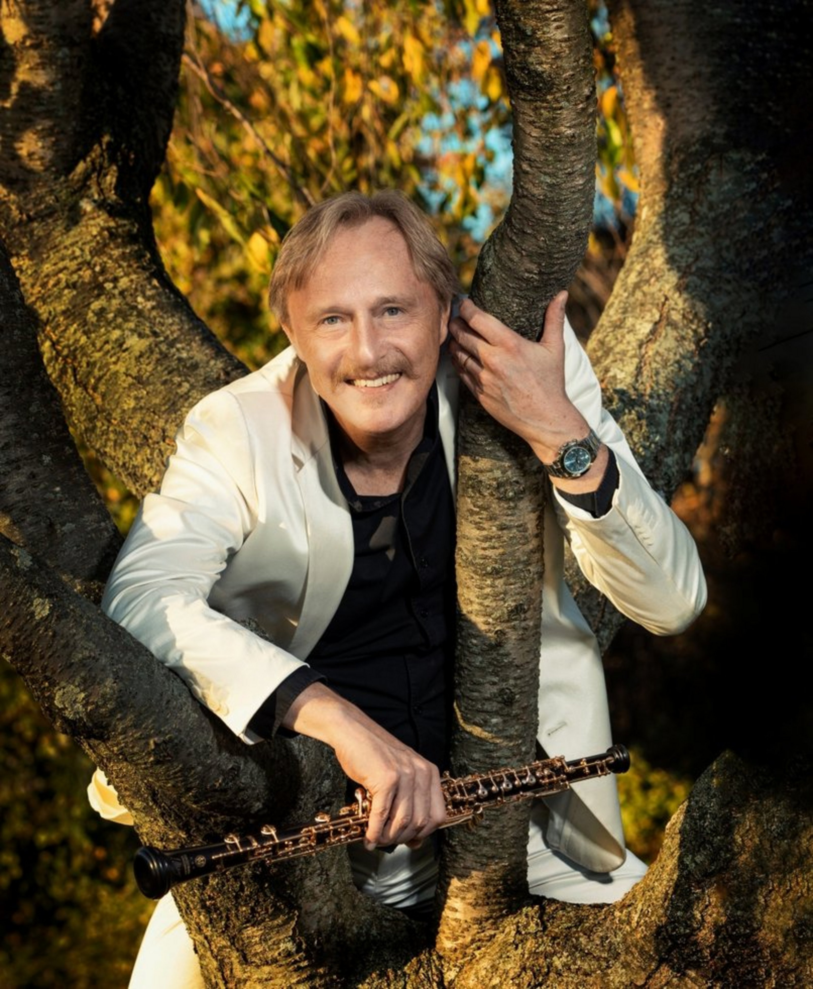 Ein Mann steht hinter einem Baum und lehnt sich durch die Äste nach vorne. In der Hand hält er eine Oboe. Er trägt eine weiße Anzugjacke und hat einen Oberlippenbart.
