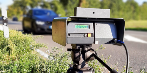 Ein Tempomessgerät steht am Straßenrand und beobachtet den Verkehr. Ein Auto fährt in Richtung des Geräts.