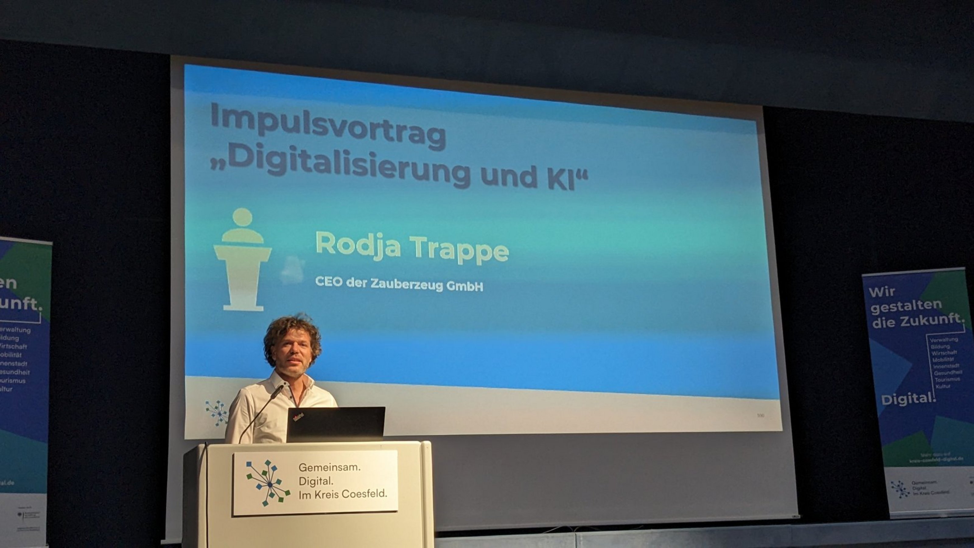 Eine Person steht an einem Rednerpult vor einer großen Präsentationsfolie. Auf der Folie steht: ‚Impulsvortrag „Digitalisierung und KI“ – Rodja Trappe, CEO der Zauberzeug GmbH‘. Auf dem Pult ist das Logo ‚Gemeinsam. Digital. Im Kreis Coesfeld.‘ zu sehen.