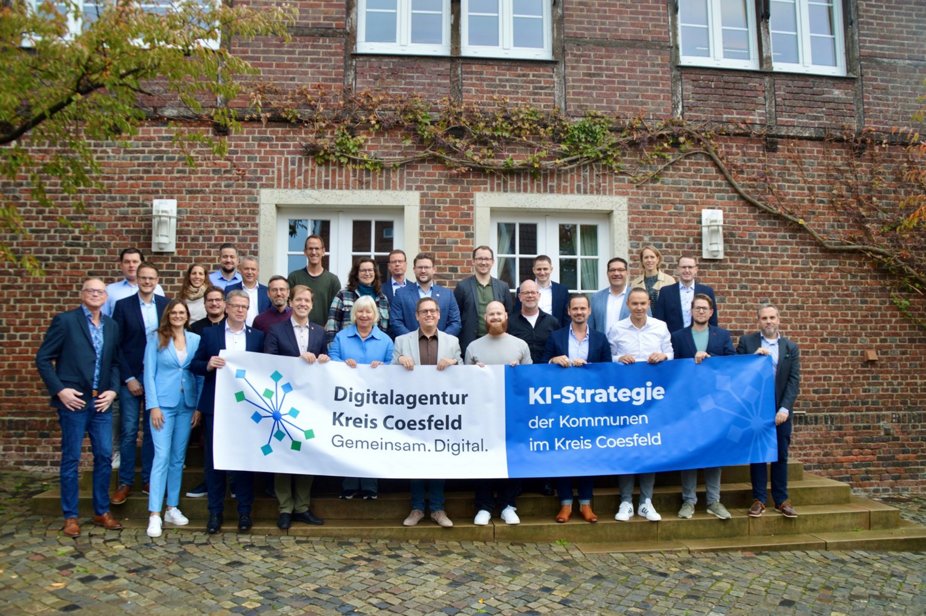 „Gruppe von Teilnehmenden vor einem Backsteingebäude hält ein Banner mit der Aufschrift ‚Digitalagentur Kreis Coesfeld Digital – Gemeinsam. Digital. Im Kreis Coesfeld.‘ und ‚KI‑Strategie‘“