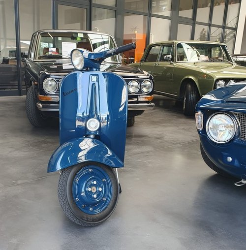 Eine klassische, blau lackierte Vespa steht im Vordergrund eines Ausstellungsraums mit mehreren Oldtimern im Hintergrund, darunter zwei Limousinen in Schwarz und Olivgrün sowie ein blauer Sportwagen. 