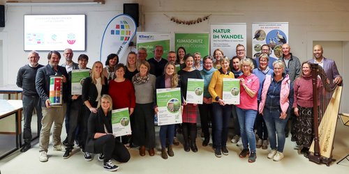 Eine große Gruppe Frauen und Männer steht zusammen in einem Raum. Einige Personen halten eine große weiß grüne Urkunde in der Hand.