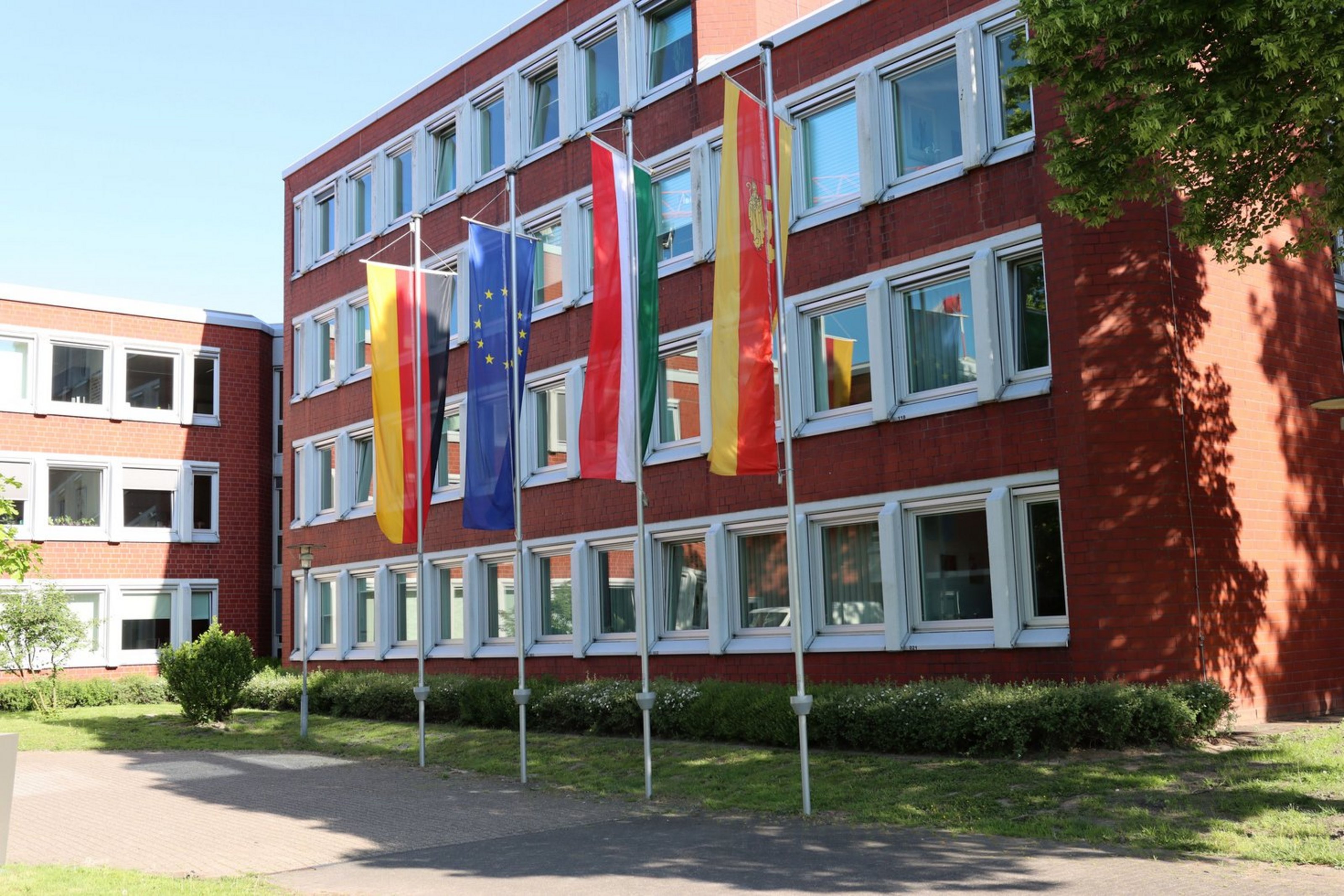 Beflaggung am Kreishaus anlässlich des 80. Jahrestages des Endes des Zweiten Weltkrieges in Europa und der Befreiung vom Nationalsozialismus (Foto: Kreis Coesfeld) Mehrstöckiges rotes Backsteingebäude mit vielen weißen Fenstern. Vor dem Gebäude stehen vier Flaggenmasten mit wehenden Flaggen: von links nach rechts die Flagge Deutschlands, der Europäischen Union, Nordrhein-Westfalens und des Kreises Coesfeld. Es ist ein sonniger Tag mit blauem Himmel, und auf dem Rasen vor dem Gebäude werfen Bäume Schatten.