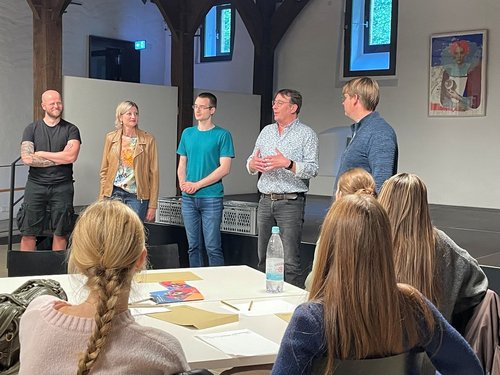 Fünf Personen stehen nebeneinander und blicken nach links. Im Vordergrund sitzen vier Personen an einem Gruppentisch und blicken zu den stehenden Personen.