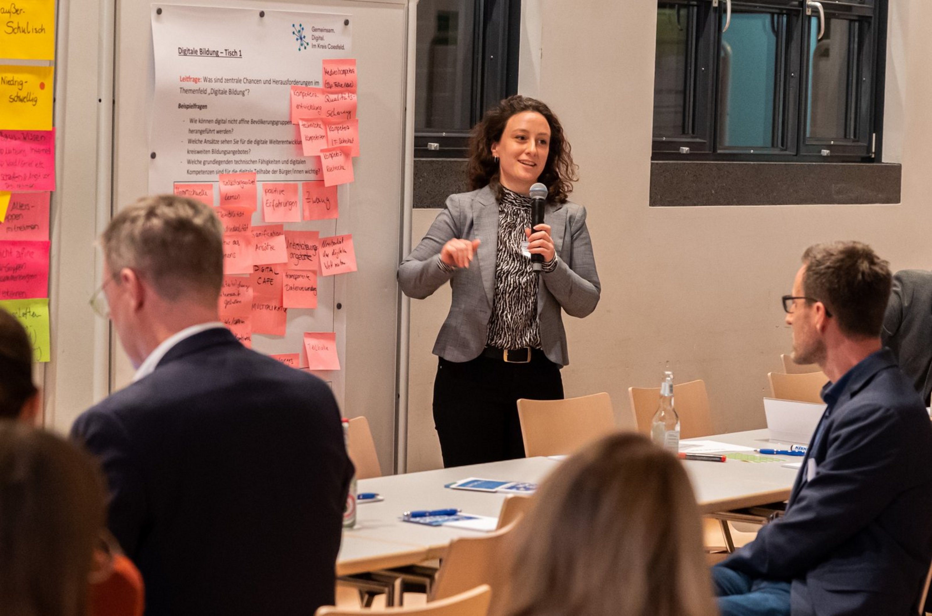 Frau steht vor einem Whiteboard mit vielen rosa Haftnotizen und spricht zu einer Gruppe von sitzenden Personen