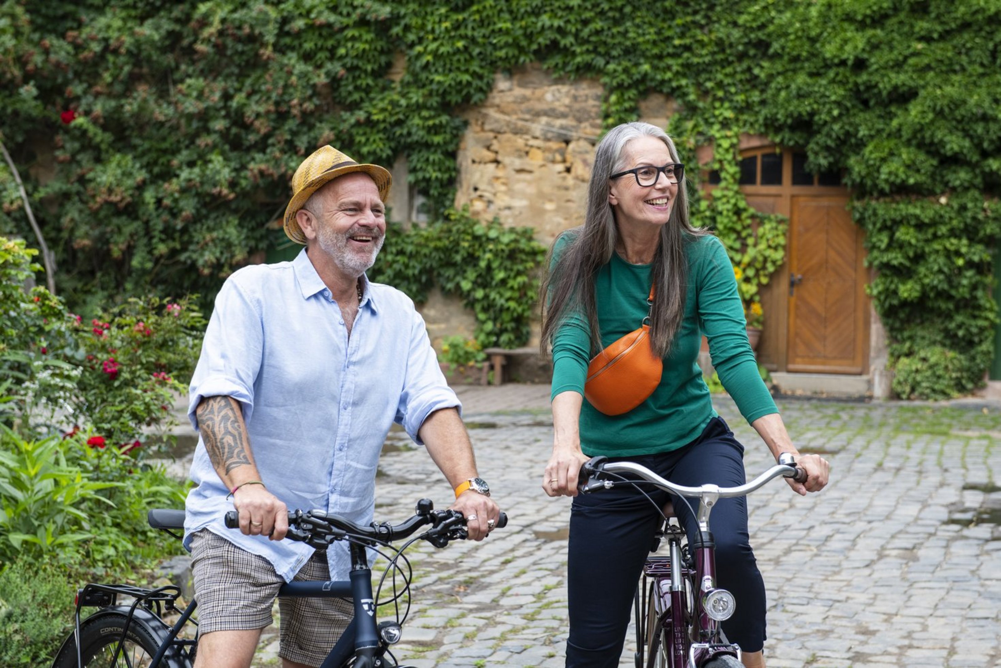 Eine Mann (links) und eine Frau (rechts) sitzen auf einem Fhrrad auf einem gepflasterten Hof vor einem Steingebäude, das von grünen Pflanzen bedeckt ist. Der Mann trägt ein Hemd und eine kurze Hose und einen Hut, die Frau trägt ein langärmeliges Shirt und eine dunkle Hose und eine Brille sowie eine Umhängetasche.