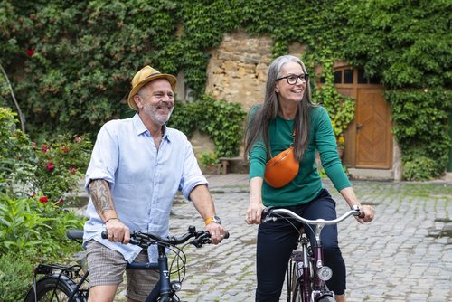 Eine Mann (links) und eine Frau (rechts) sitzen auf einem Fhrrad auf einem gepflasterten Hof vor einem Steingebäude, das von grünen Pflanzen bedeckt ist. Der Mann trägt ein Hemd und eine kurze Hose und einen Hut, die Frau trägt ein langärmeliges Shirt und eine dunkle Hose und eine Brille sowie eine Umhängetasche.
