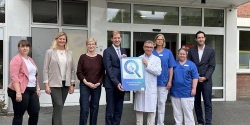 Acht Männer und Frauen stehen vor dem Eingangsbereich eines Gebäudes. Zwei der Männer halten gemeinsam ein weiß-blaues Schild auf dem Hygiene und Prävention steht.
