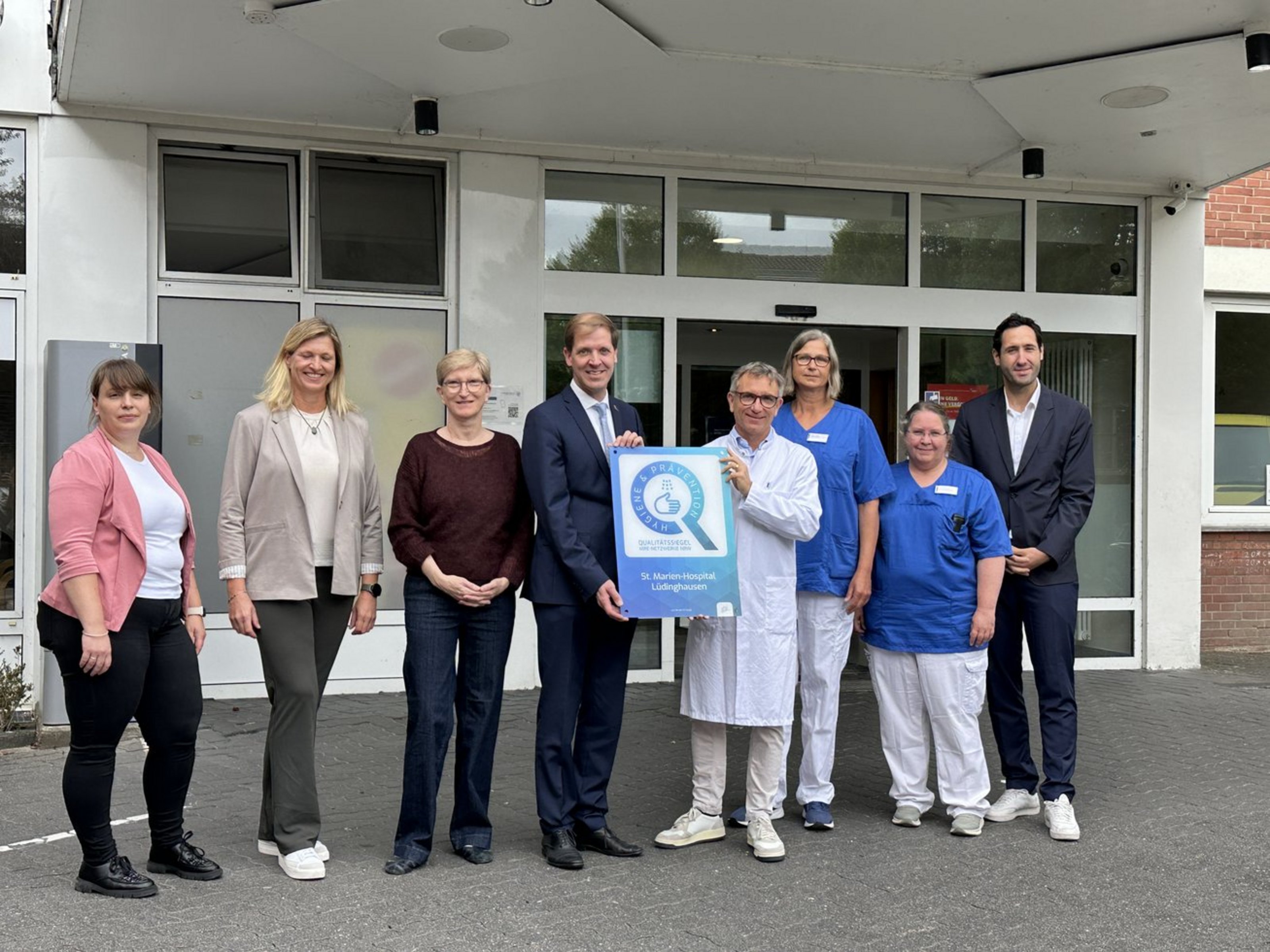 Acht Männer und Frauen stehen vor dem Eingangsbereich eines Gebäudes. Zwei der Männer halten gemeinsam ein weiß-blaues Schild auf dem Hygiene und Prävention steht.