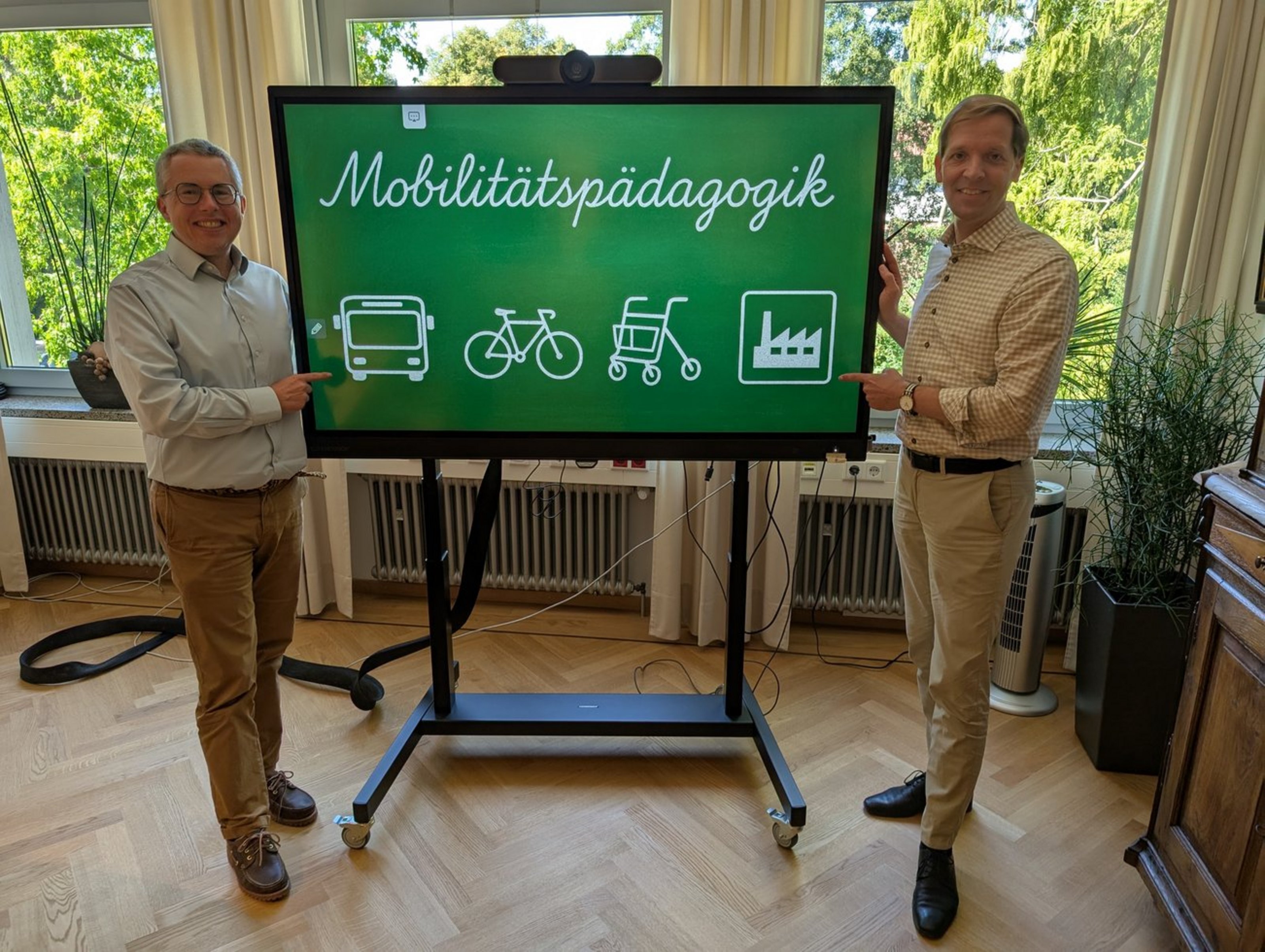 Der Kreis Coesfeld möchte zielgruppenorientiertes Mobilitätsmanagement voranbringen (Foto: Kreis Coesfeld). Zwei Männer stehen in einem hellen Raum neben einem großen Bildschirm auf einem Rollständer. Auf dem grünen Bildschirm steht in weißer Schrift „Mobilitätspädagogik“.