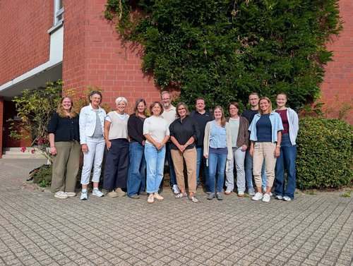 Dreizehn Männer und Frauen stehen vor einem roten Gebäude und einem Baum und lächeln in die Kamera.
