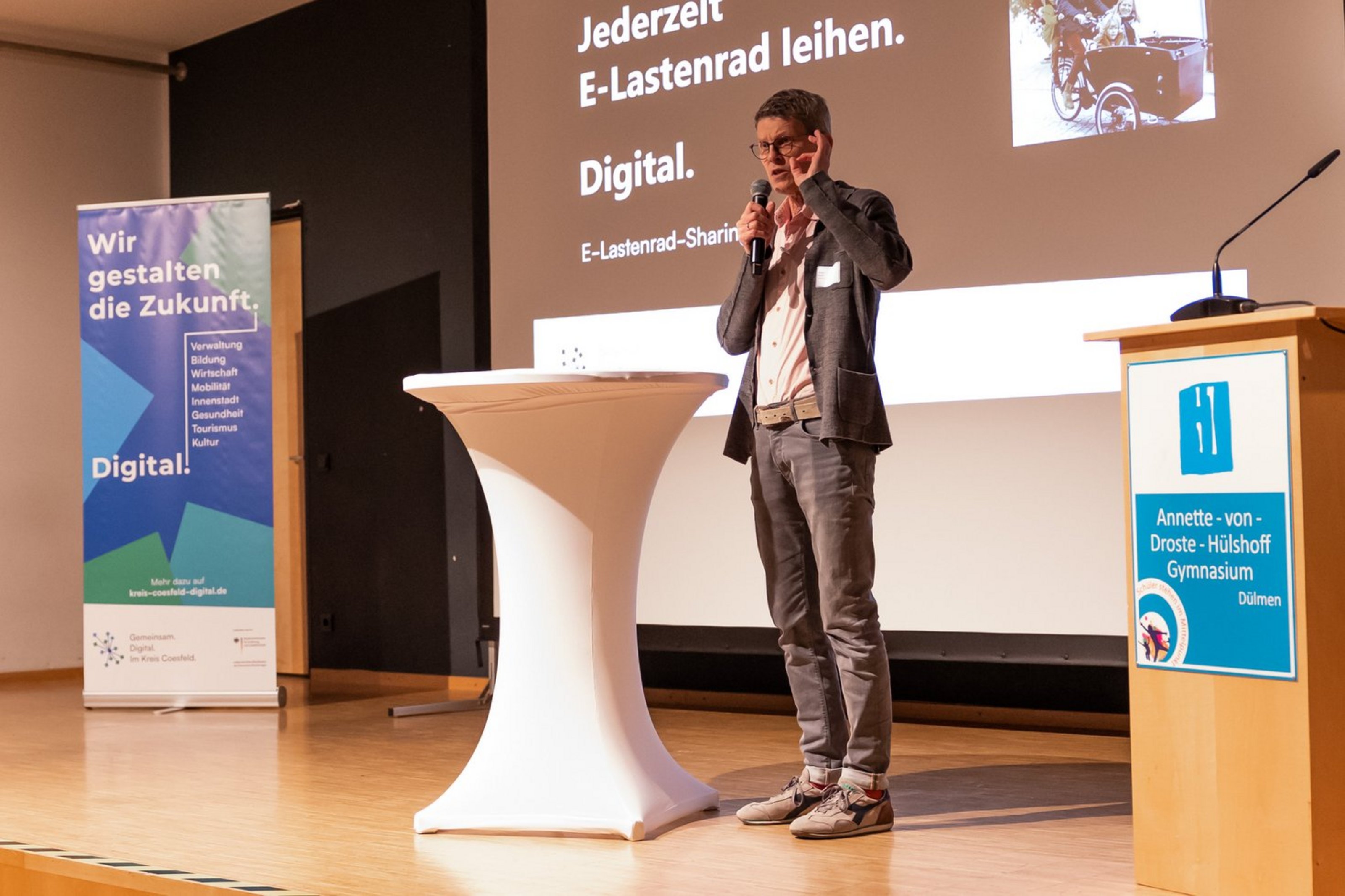 Mann steht auf Bühne neben Stehtisch und spricht vor Publikum, im Hintergrund zwei Banner mit Text und Bild